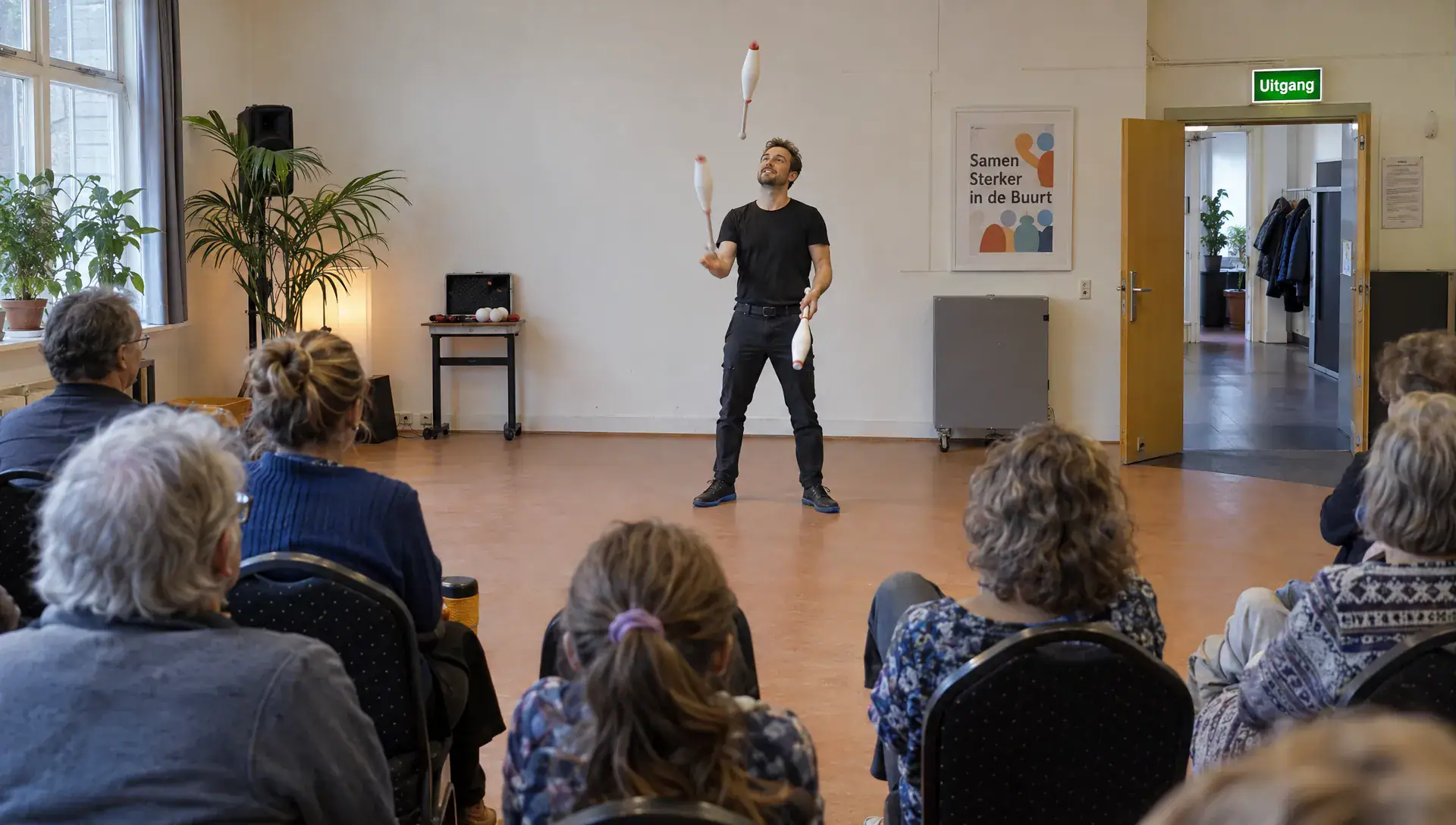 Een jongleur treedt op voor zittende bezoekers in een buurthuis, met een vrij speelvlak en zichtbare doorgang langs de zaalwand.